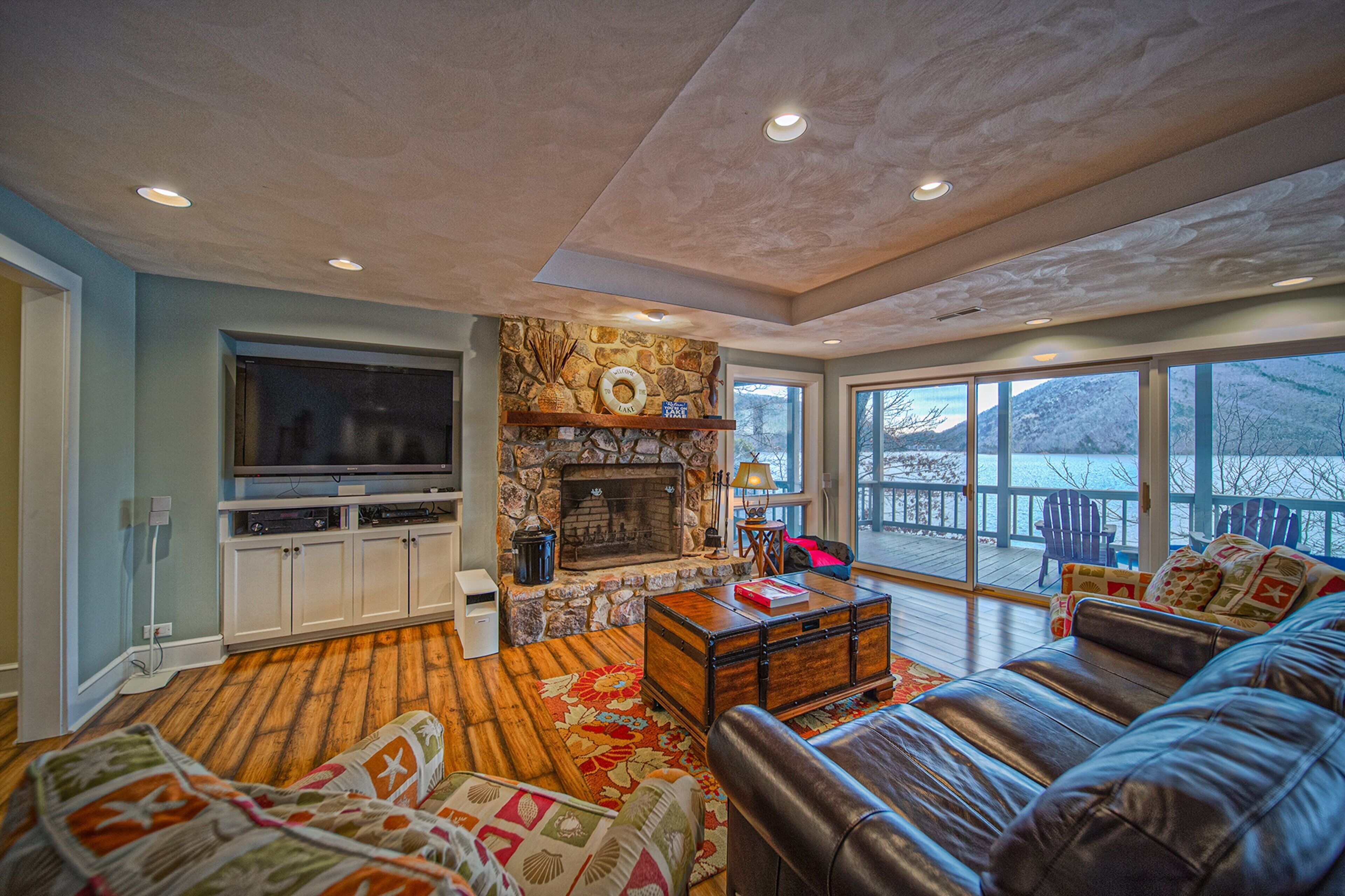 A living room with a stone fireplace, a television, leather sofa, and a large window overlooking a deck.