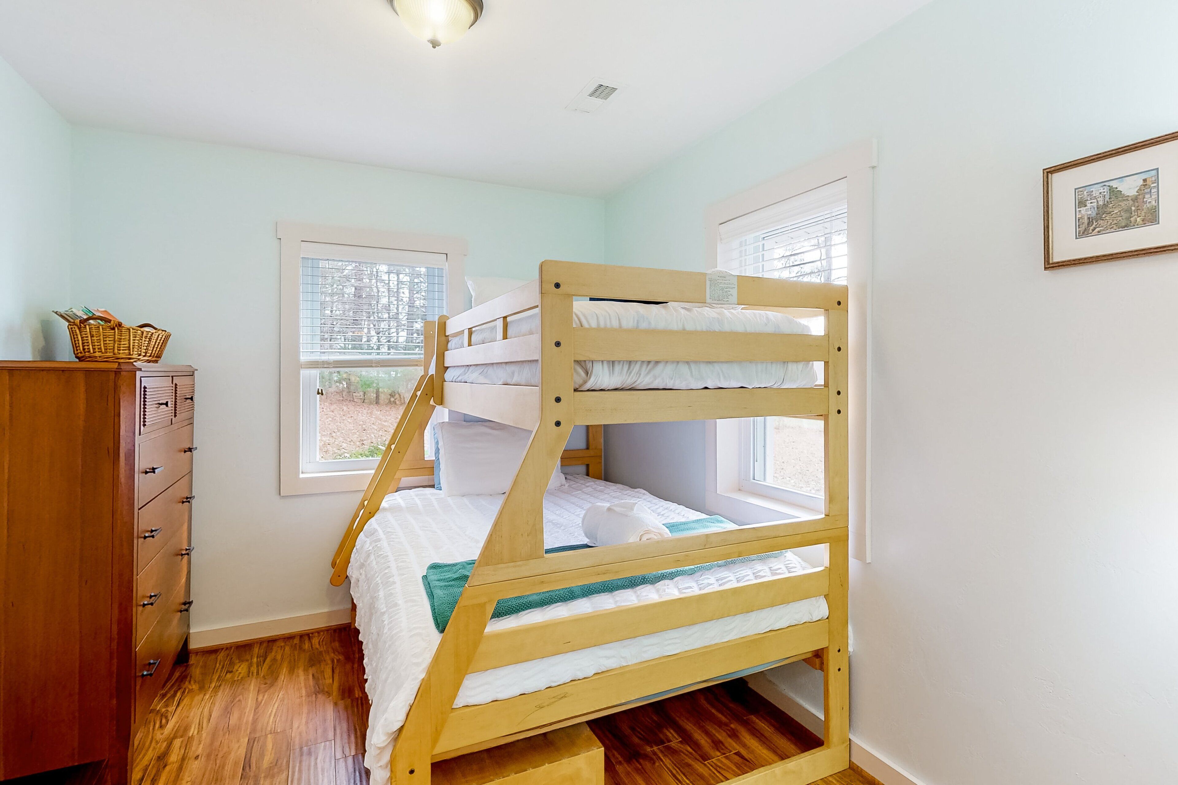 A bunk bed in a room with a wooden dresser, a window with blinds, and a framed picture on the wall.