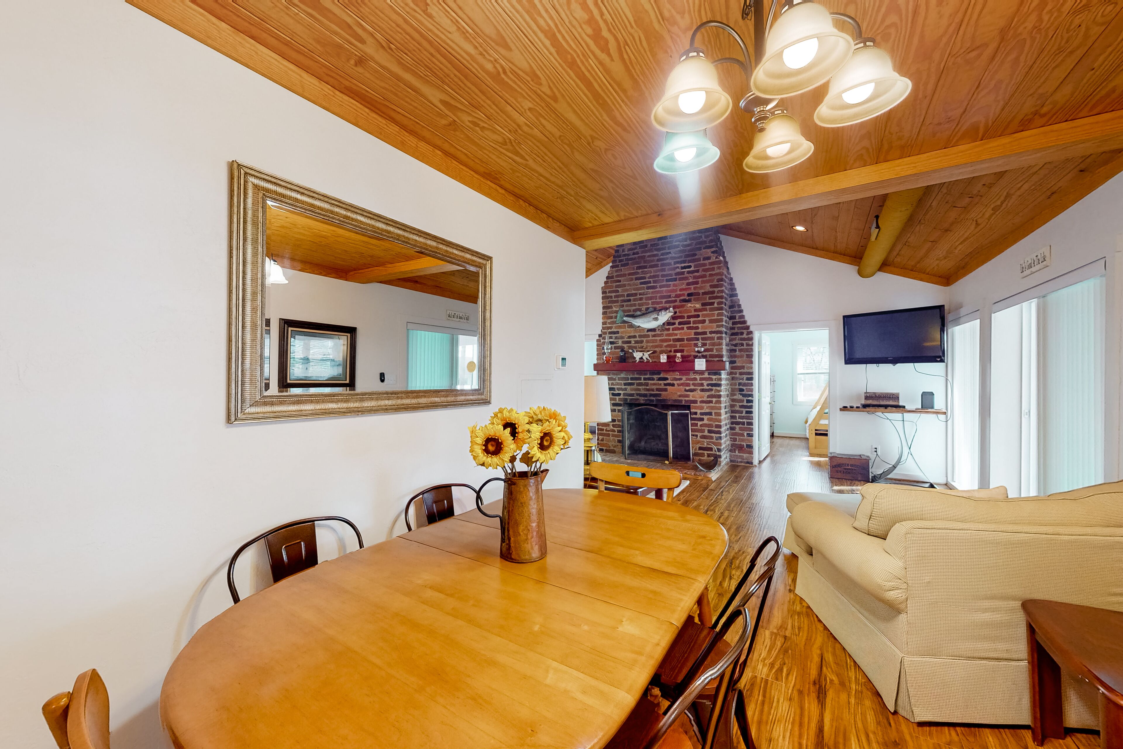 Un comedor con una mesa de madera, sillas y un jarrón con girasoles. Se observa una chimenea al fondo y un televisor montado en la pared.