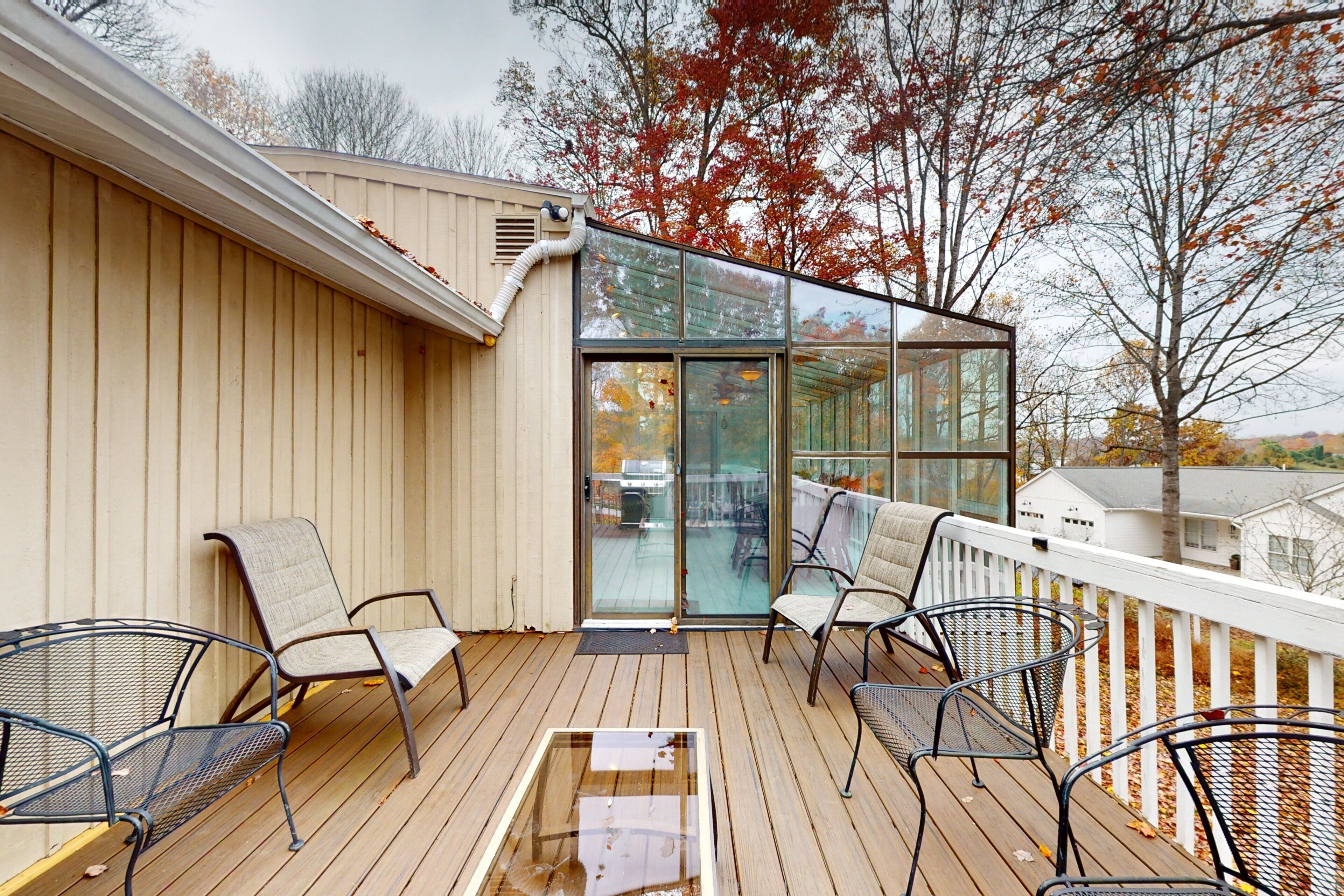 Une terrasse en bois avec des chaises en métal, une porte vitrée donnant sur un espace abrité et une rambarde blanche.