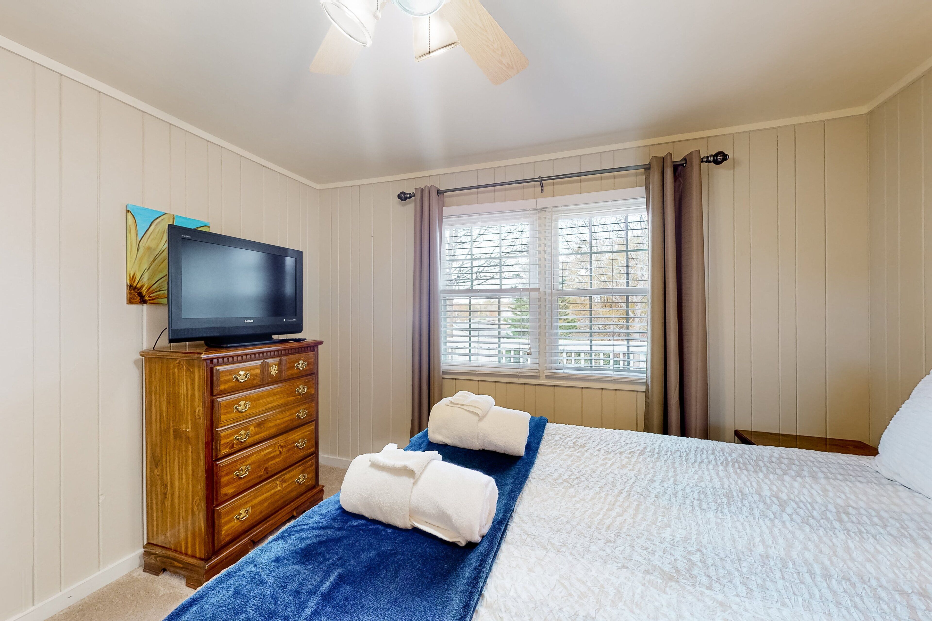 A hotel room with a bed, a television on a wooden dresser, a window with curtains, and a painting on the wall.