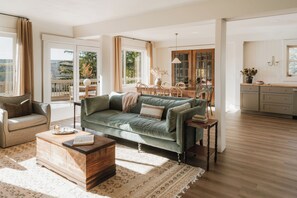 A spacious living room with a green sofa, a wooden coffee table, and a dining area with a wooden table and chairs. - Private Cabin in the Woods: Hot Tub, Pool, Sauna (Skaneateles)