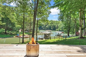 Una terraza de madera con un pozo de fuego, rodeada de árboles y vistas a un lago.