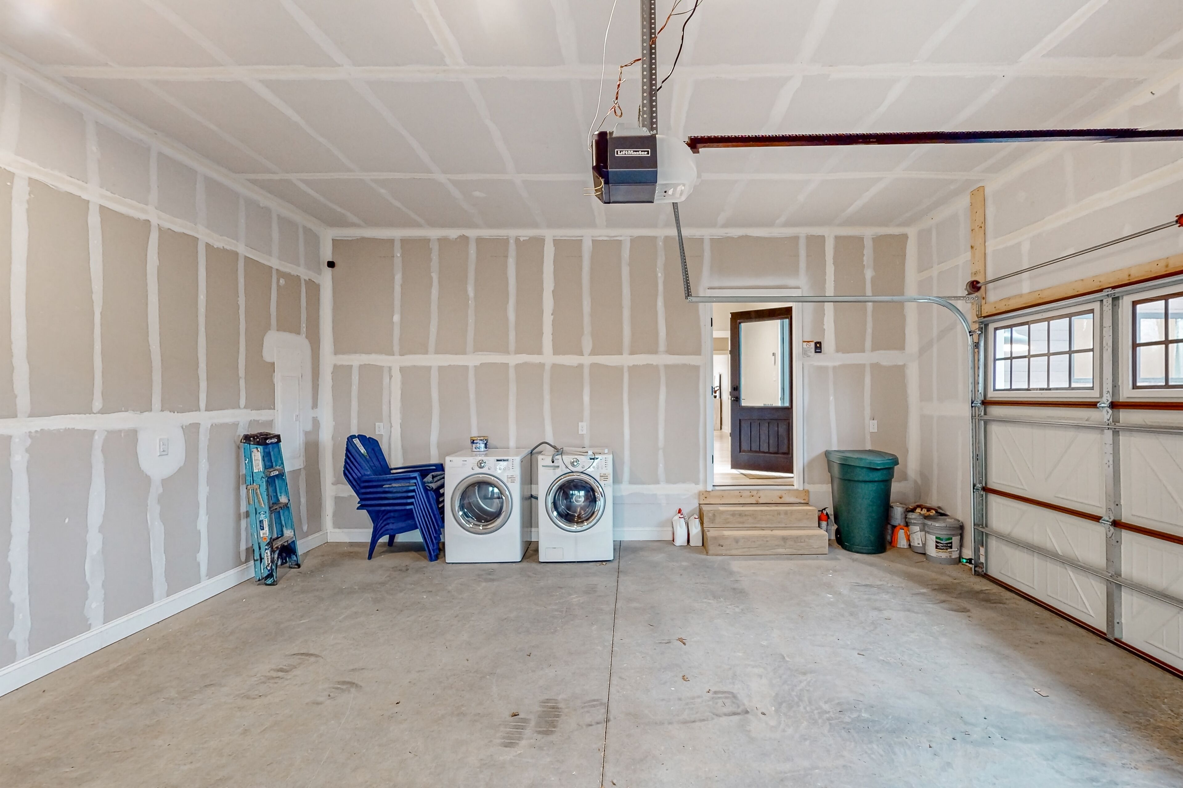 A garage with two stacked washing machines, a ladder, and a trash bin.