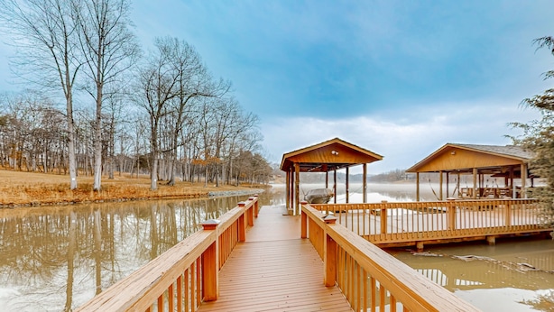 A wooden pier leading to a covered area with a view of a calm lake and trees. - Lawi Lakehouse - Stunning Lakefront Oasis With a Kayak, Large Deck, Games, & Tranquil Views (Wirtz)