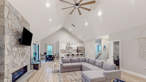 A spacious living room with a stone fireplace, a large sectional sofa, and a ceiling fan. - Lawi Lakehouse - Stunning Lakefront Oasis With a Kayak, Large Deck, Games, & Tranquil Views (Wirtz)
