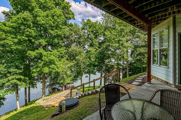 A covered outdoor area with a view of a lake, a wooden deck, and a glass table and chair. - Still Waters - Lakefront Luxury Home With Views, Dock, Home Theater & Game Room (Goodview)