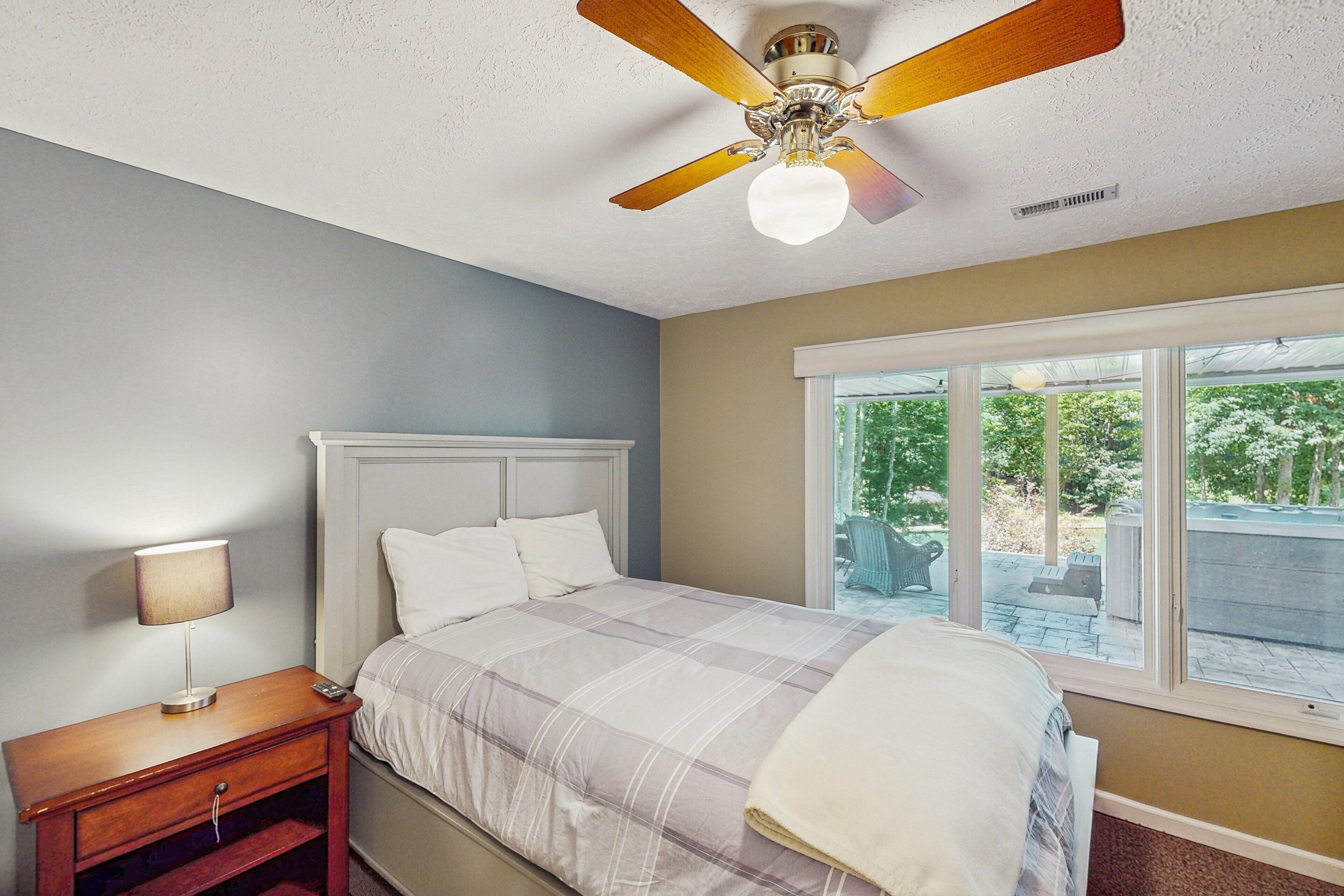 A bedroom with a bed, a nightstand, a ceiling fan, and a window with a view of greenery.