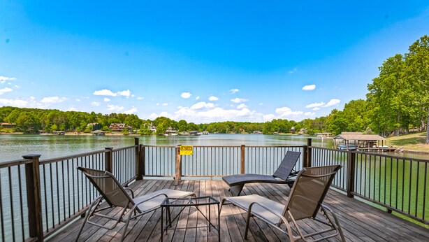 A wooden deck with two chairs and a table, overlooking a lake with houses in the background. - Cottage at the Lake - Waterfront Lake-view Home With Dock, Fireplace, Pool Table, Kayaks, & Firepit (Moneta)