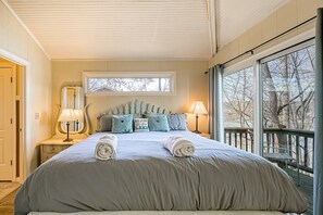 A bedroom with a large bed, two bedside tables with lamps, a window with a view of trees, and a door to the left.
