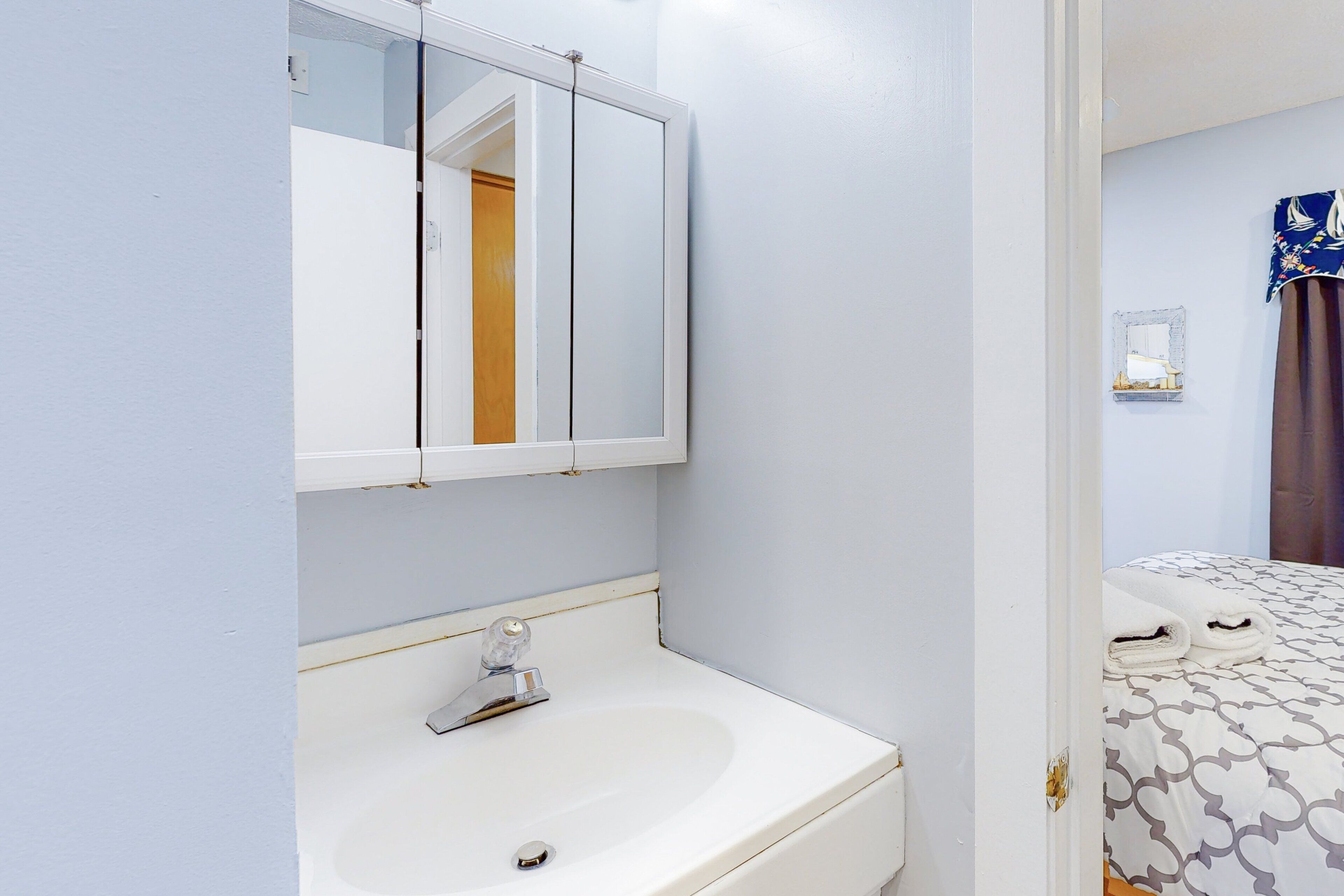 A bathroom with a sink, mirror, and a door leading to a bedroom with a bed and curtains.