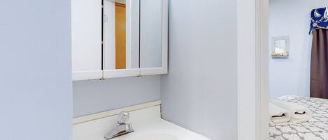 A bathroom with a sink, mirror, and a door leading to a bedroom with a bed and curtains.