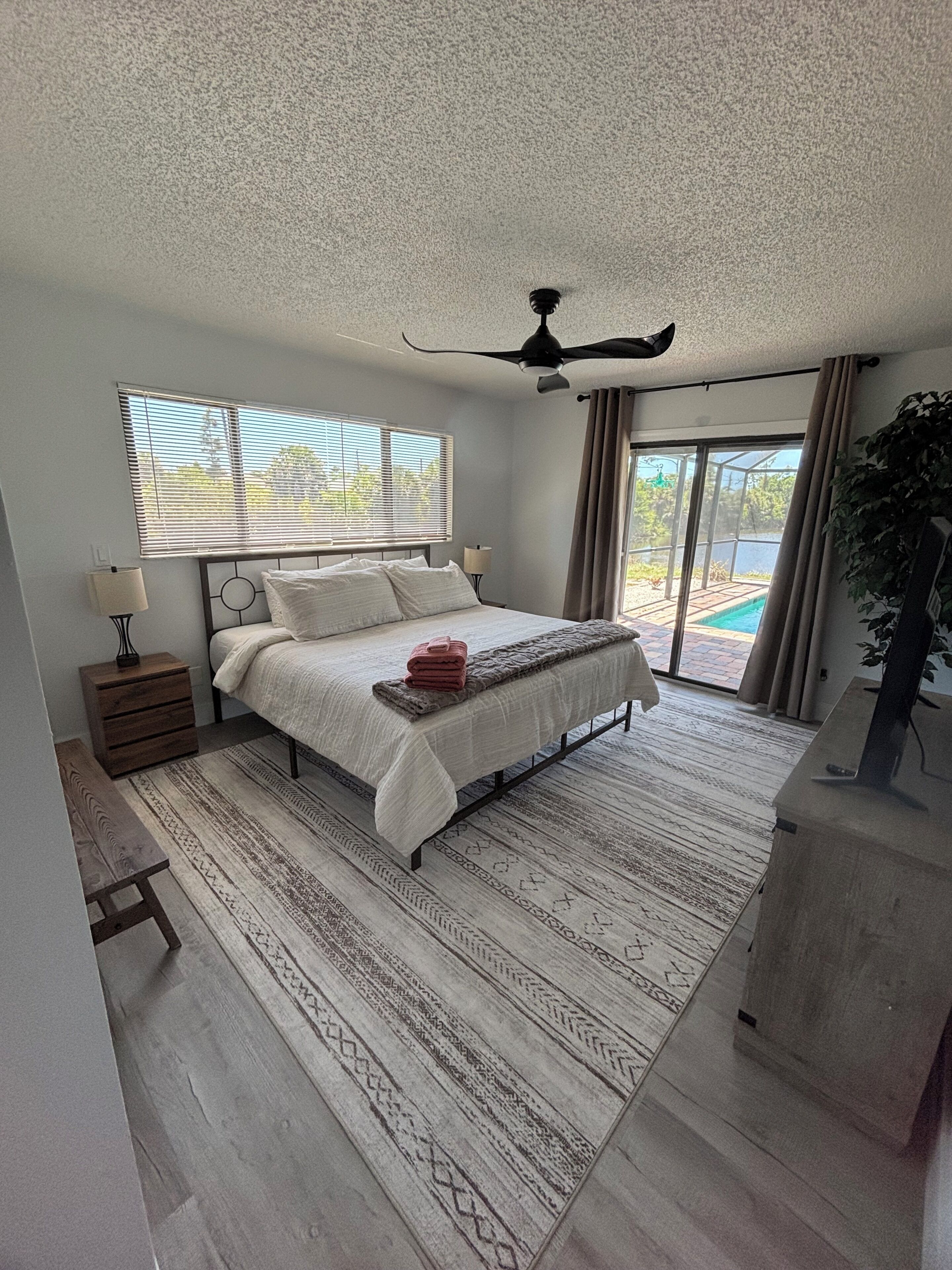 A bedroom with a bed, a ceiling fan, a window with blinds, a bedside table, a nightstand, a bench, a rug, and a view of a swimming pool.