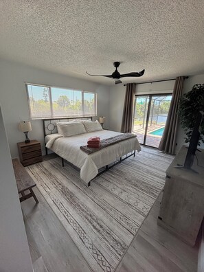 Ein Schlafzimmer mit Bett, Deckenventilator, Fenster mit Jalousien, Nachttisch, Sitzbank, Teppich und Blick auf einen Swimmingpool.