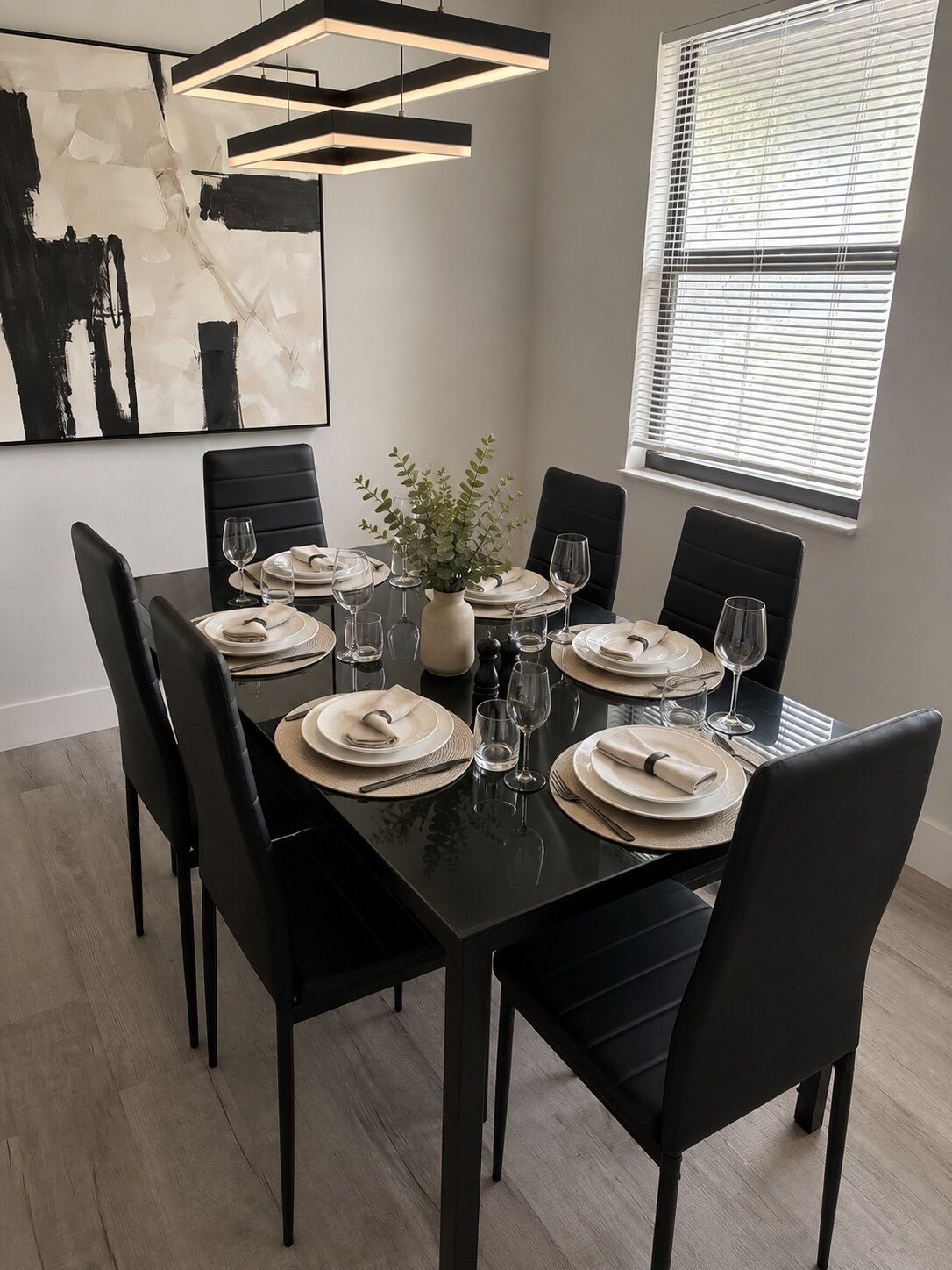 Comedor con una mesa rectangular dispuesta para cuatro, sillas negras, un jarrón con follaje y una lámpara colgante moderna.