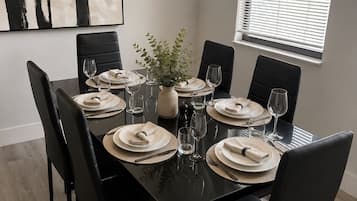 Comedor con una mesa rectangular dispuesta para cuatro, sillas negras, un jarrón con follaje y una lámpara colgante moderna.