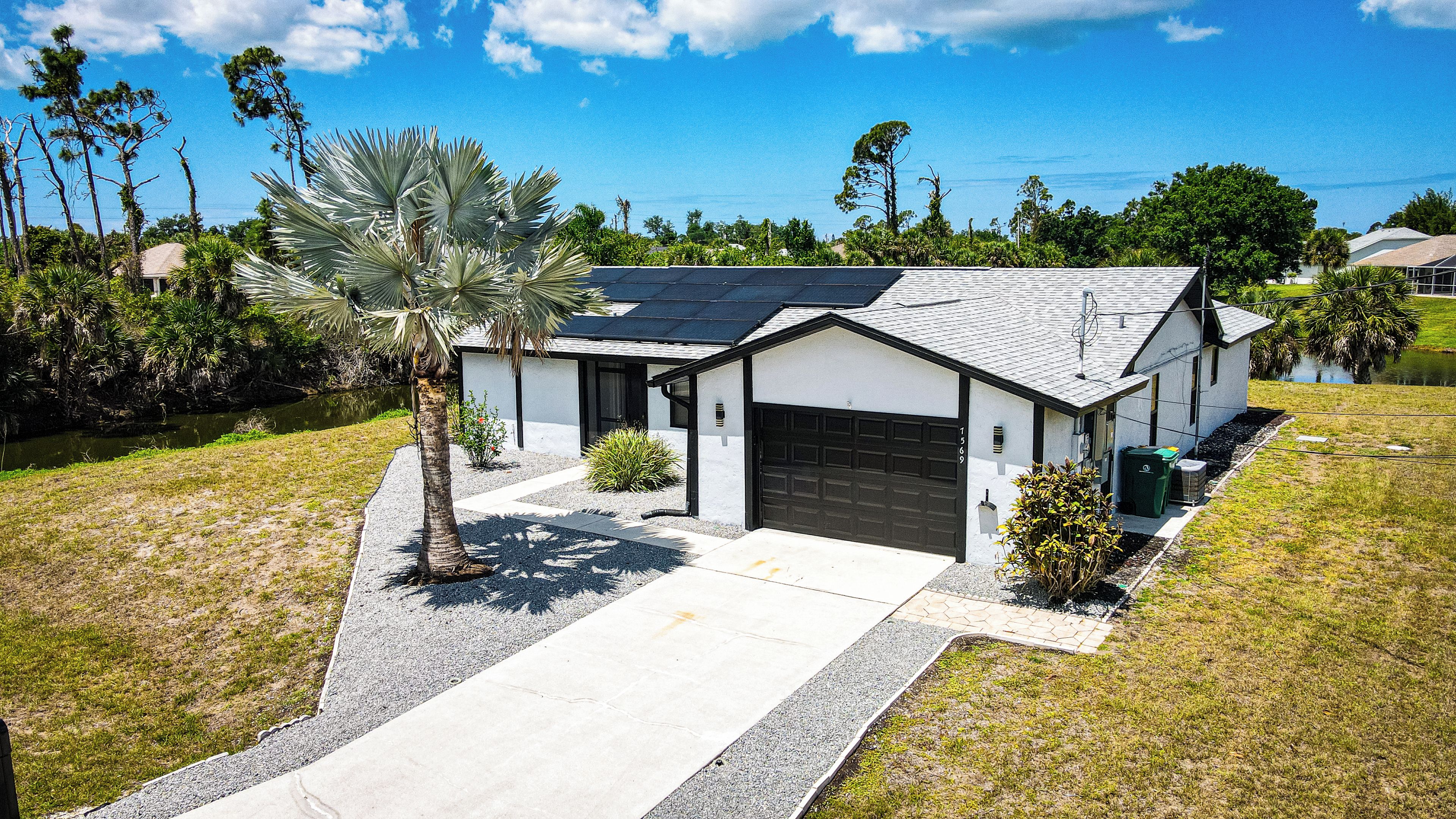 A single-story house with a garage, a solar panel roof, and a small pond in the background.