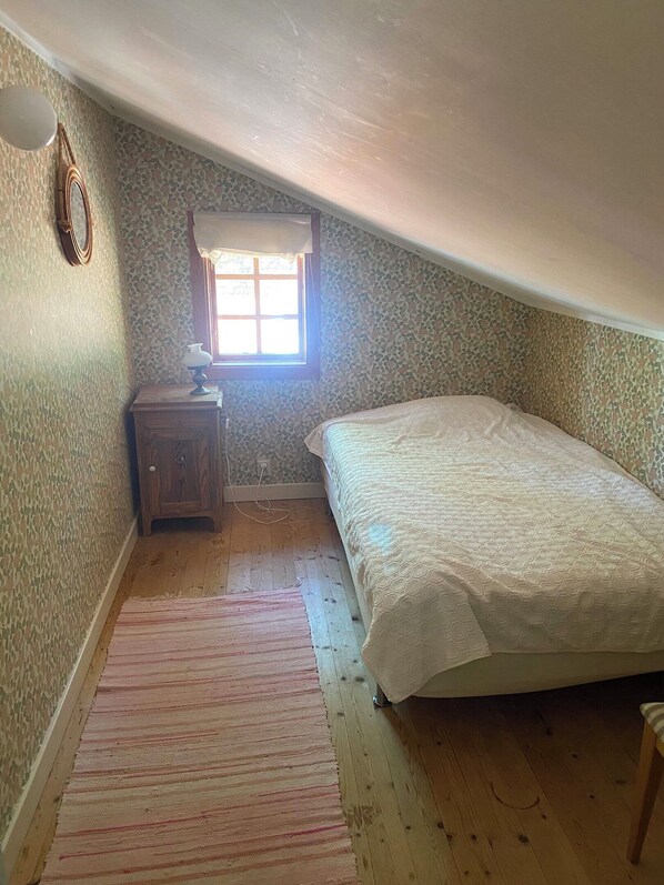 Une chambre mansardée avec un plafond en pente, un lit simple, une table de chevet, une petite fenêtre et un mur orné d’un motif.