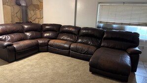 A brown leather sectional sofa in a living room with a stone wall, a window with blinds, and a carpeted floor.
