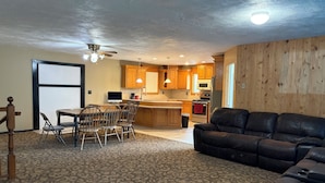 A living room with a sofa, a dining area with chairs and a table, a kitchen with wooden cabinets and appliances, and a ceiling fan.