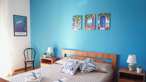 Cama bem arrumada com almofadas, uma cadeira, mesa de cabeceira com abajur e obras de arte na parede.