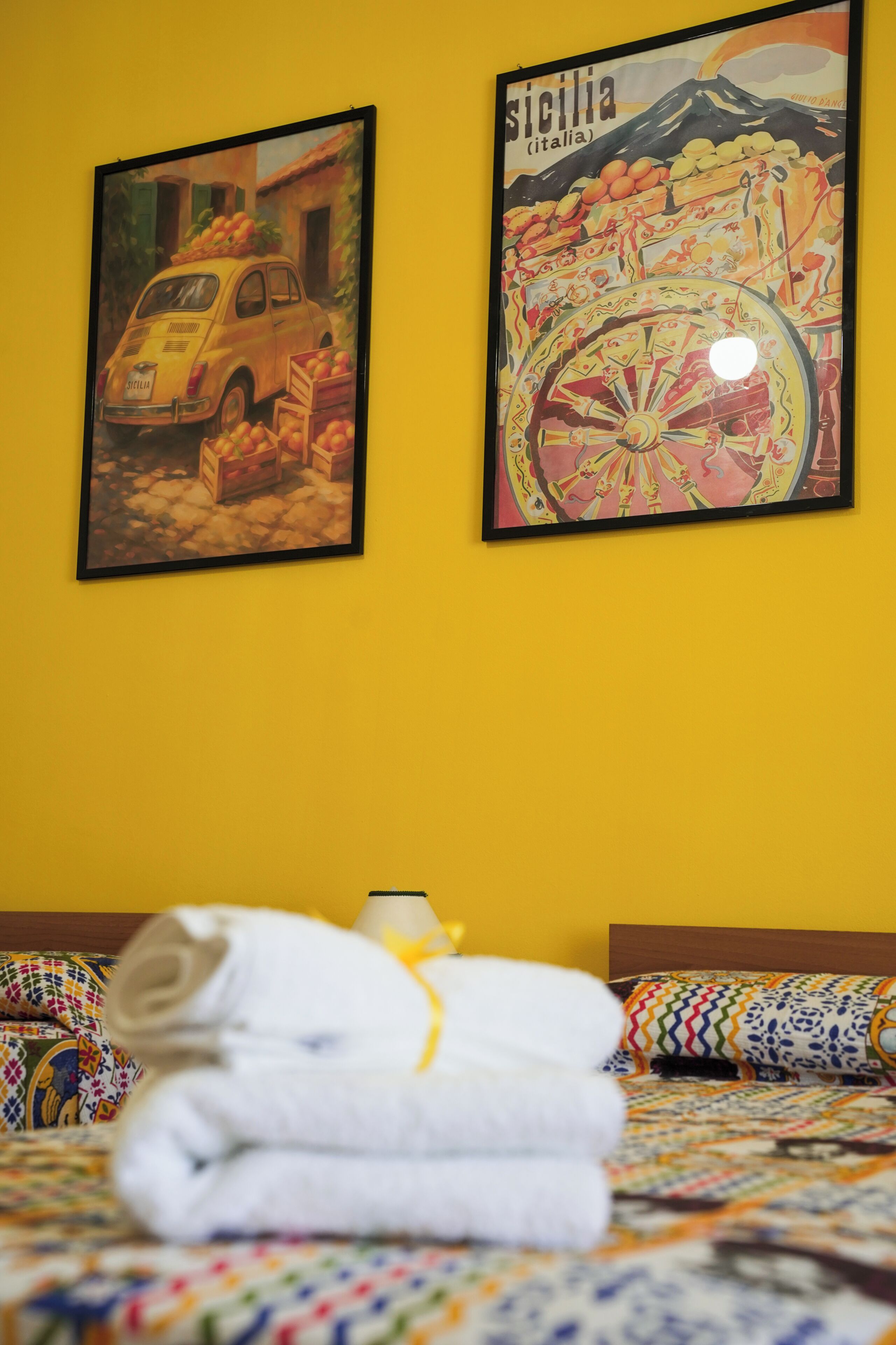 A bed with a colorful quilt, a rolled towel, and two framed pictures on a yellow wall.