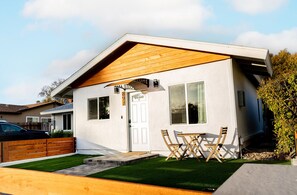 Ein modernes eingeschossiges Haus mit weißer Fassade, einem Holzdach und einem kleinen Vorgarten mit Rasen und einem Patio-Tisch.