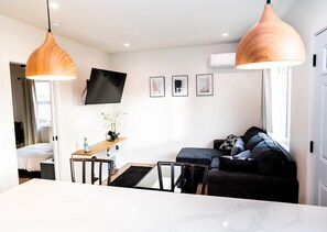 Ein modernes Wohnzimmer mit einer Couch, einem an der Wand befestigten Fernseher, einem Esstisch und Pendelleuchten.