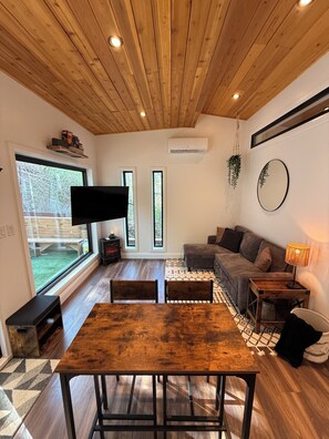 Ein modernes Wohnzimmer mit Holzboden, einem großen Fernseher, einer Couch, einem Esstisch und Blick ins Freie.