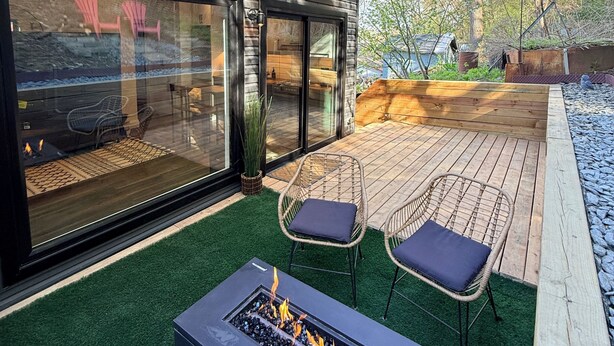 Een modern terras buiten met een vuurkuil, twee rieten stoelen en een houten terras.