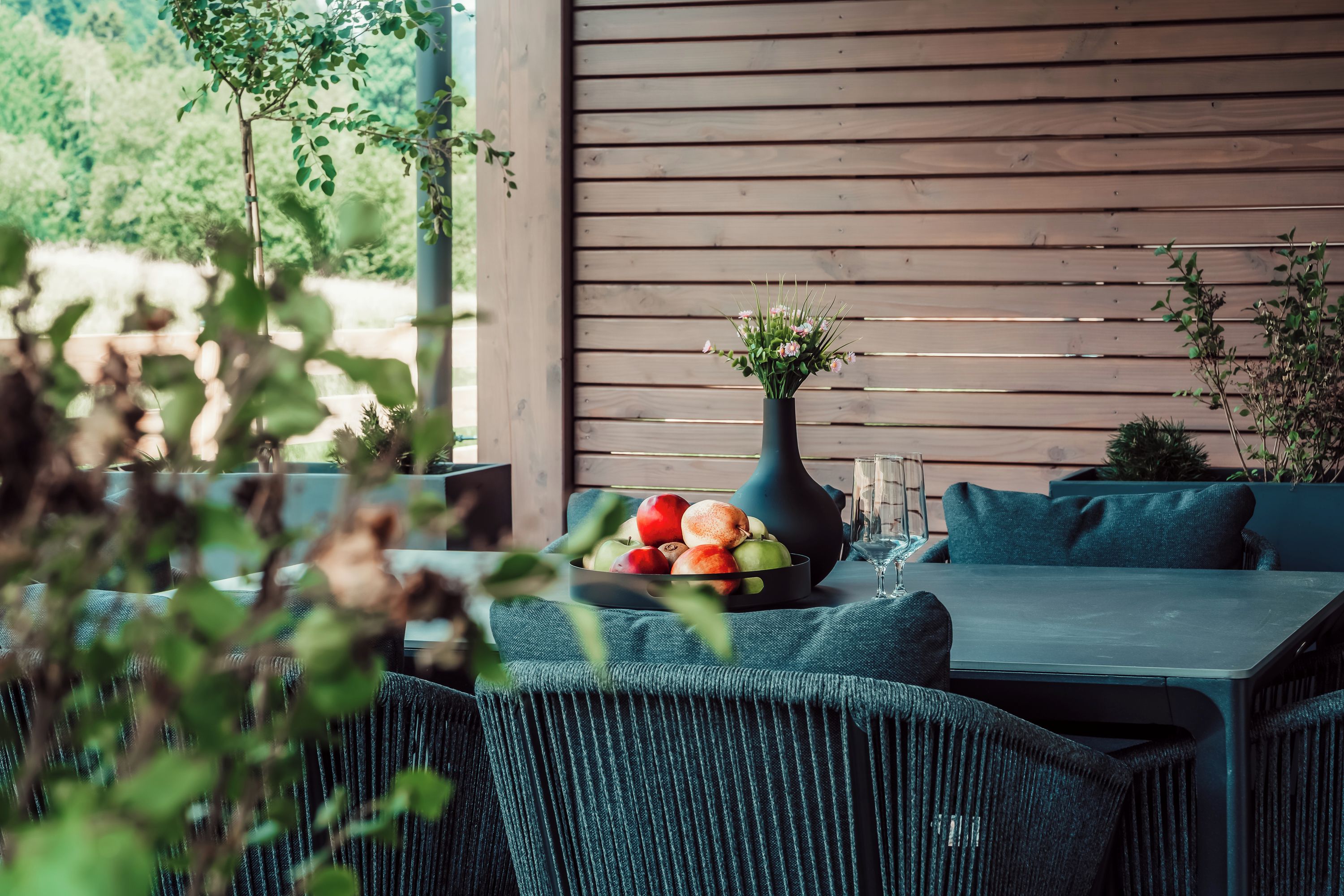 An outdoor seating area with a table set with fruits, a vase with flowers, and a glass of wine.