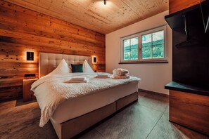 Un dormitorio con techo de madera, una cama grande con ropa blanca, una ventana con vista a árboles y un televisor a la derecha.
