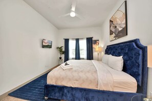 A bedroom with a bed, a ceiling fan, a painting, a window with curtains, and a nightstand with a lamp. - Relaxing Houston Getaway | 2 King Suites + Game Ro (Houston)