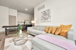 A modern living room with a sofa, a glass coffee table, and a kitchen in the background. - Relaxing Houston Getaway | 2 King Suites + Game Ro (Houston)