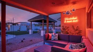 Een terras buiten met een bank, een kleine tafel en een neonbord waarop staat 'Wanderlust & desert dust'.