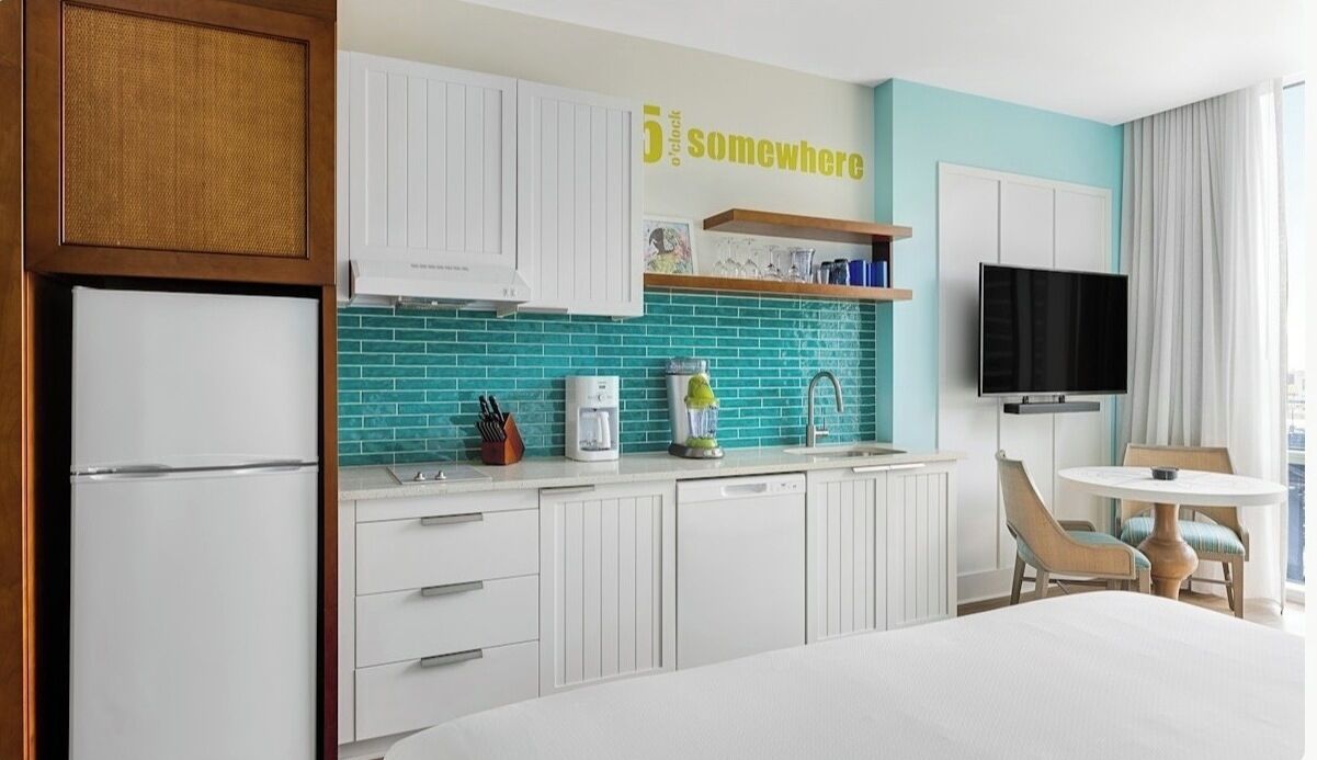 A modern kitchen with white cabinets, a teal backsplash, a coffee maker, and a TV mounted on the wall.