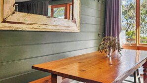 Une table en bois sur laquelle est posé un vase contenant des plantes séchées, une fenêtre donnant sur des arbres et un miroir partiellement visible accroché au mur.