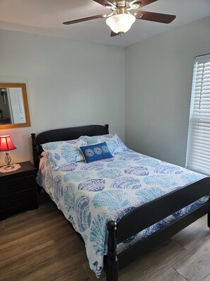 Ein Schlafzimmer mit einem Bett, einem Nachttisch, einer Lampe, einem Deckenventilator, einem Fenster mit Jalousien und einem Spiegel an der Wand.