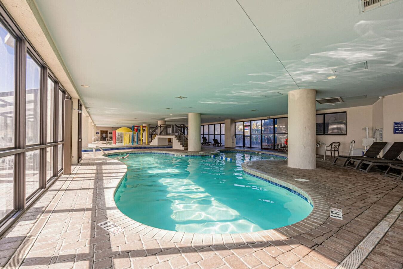 An indoor swimming pool with clear glass walls, a tiled floor, and a few lounge chairs.