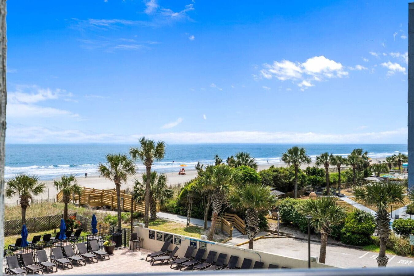 Blick auf einen Strand mit Liegestühlen, Palmen und einer Holzterrasse.