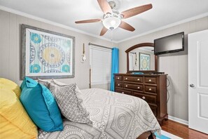 Um quarto com cama, cômoda, ventilador de teto, televisão e uma obra de arte emoldurada na parede.