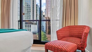 A hotel room with a bed, a red checkered chair, and a large window with a city view. - Cosy Cityview Modern Studio | Pool - Gym - Aircon (Auckland CBD)