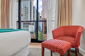 Ein Hotelzimmer mit einem Bett, einem rot karierten Sessel und einem großen Fenster mit Blick auf die Stadt.