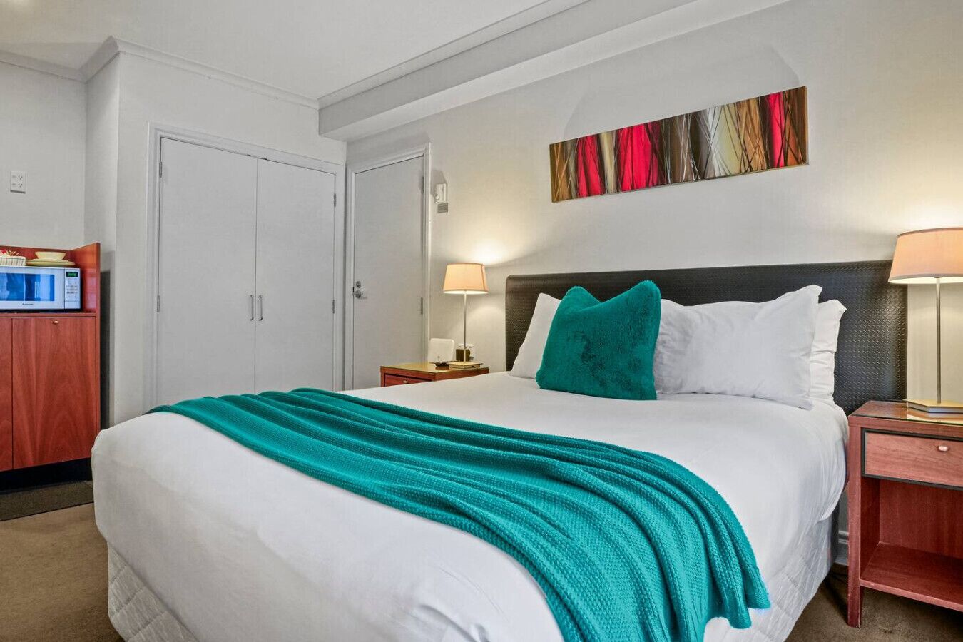 A hotel room with a large bed, a nightstand, a desk with a lamp, a wardrobe, and a painting on the wall.