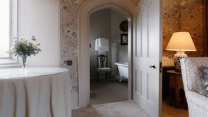 A room with a round table, a chair, a lamp, and a door leading to a bathroom with a bathtub.