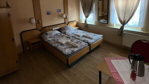 Een kamer met een bed, een tafel met waterkoker, een stoel, een kledingkast en een raam met gordijnen.