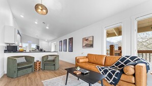 A modern living room with a brown leather sofa, a green sofa, and a black coffee table. There is a kitchen in the background with white cabinets and a hanging light fixture. - Game Room Condo Walk to The Landing with Pools and Creek Views, Family Friendly (Branson)