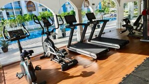 A gym with treadmills, an elliptical, and dumbbells, with a view of a pool and buildings in the background. -  Cozy 3BR Condo with AC at Negombo Santorini Resort, Close to Airport (Negombo)