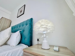 Una cama bien hecha con ropa de cama blanca, cabecera color turquesa, lámpara de plumas blancas y una mesita de noche con control remoto.