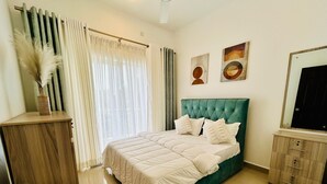 A bedroom with a green upholstered bed, white bedding, a wooden nightstand, a mirror, and a green rug. -  Cozy 3BR Condo with AC at Negombo Santorini Resort, Close to Airport (Negombo)